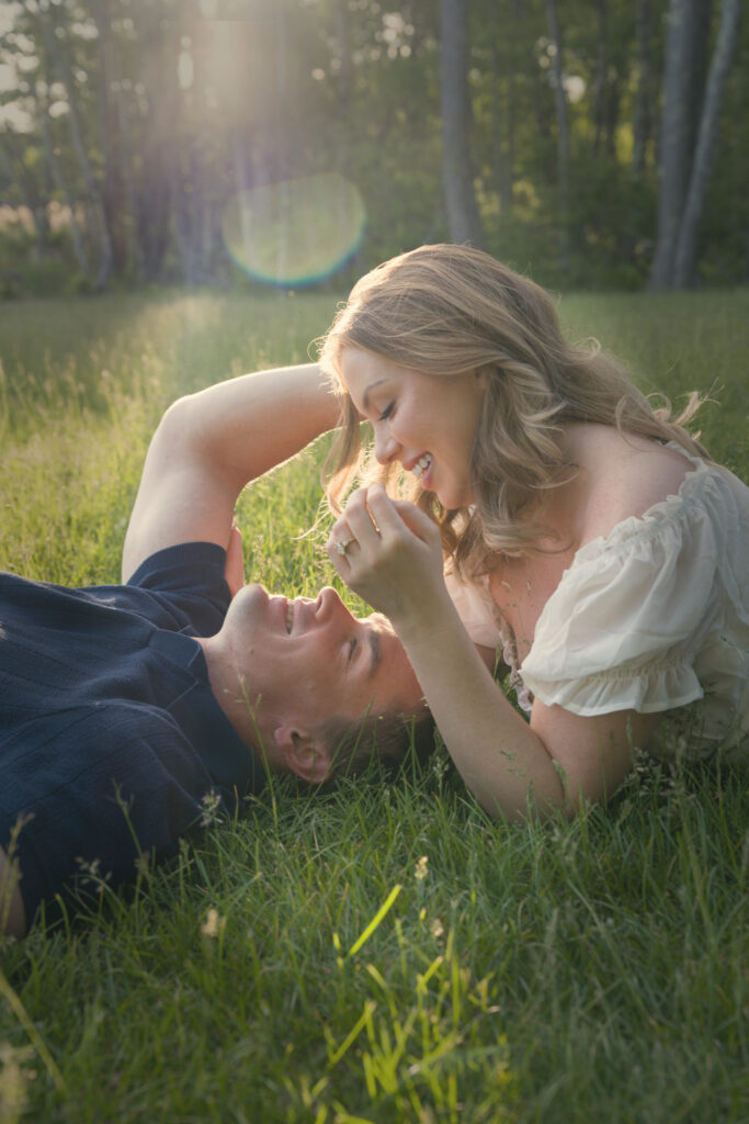 Engaged couple lying together in tall grass with the sun flaring through the trees