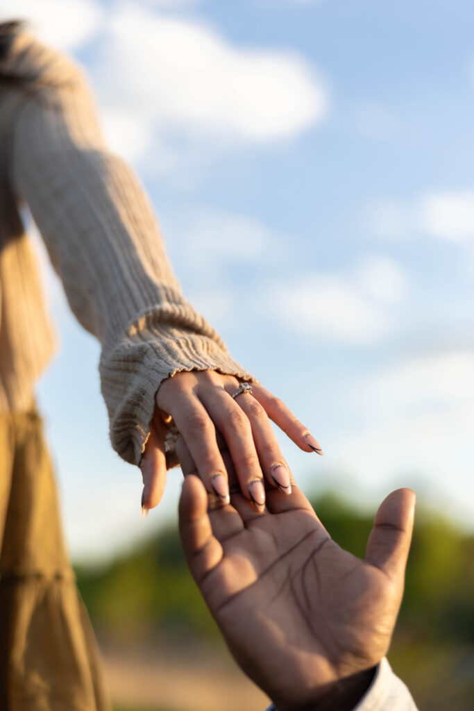 Bride's hand with her engagement ring reaching toward her partner's open hand against the sky